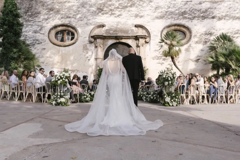 The bride walking towards the ceremony, guests seated before the stone façade, Jardines de Alfabia, Mallorca